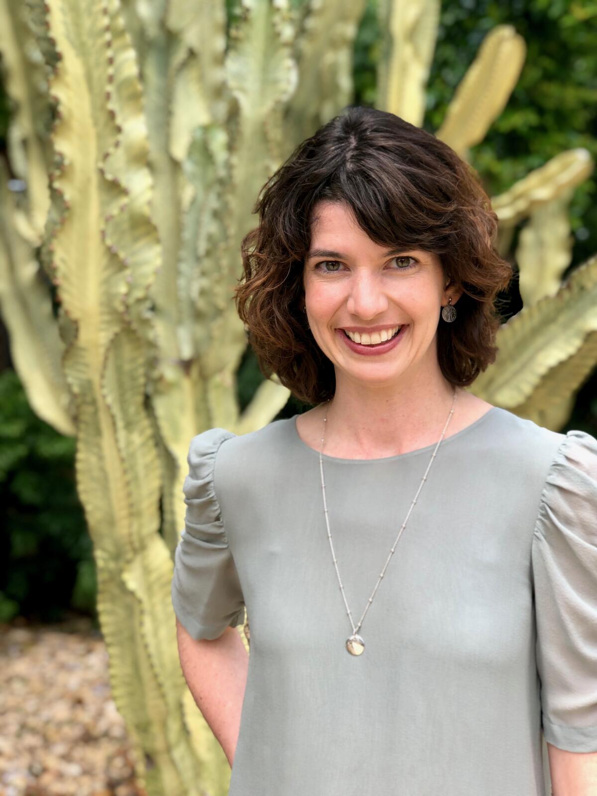 A woman in a gray blouse smiles. A cactus is in the background