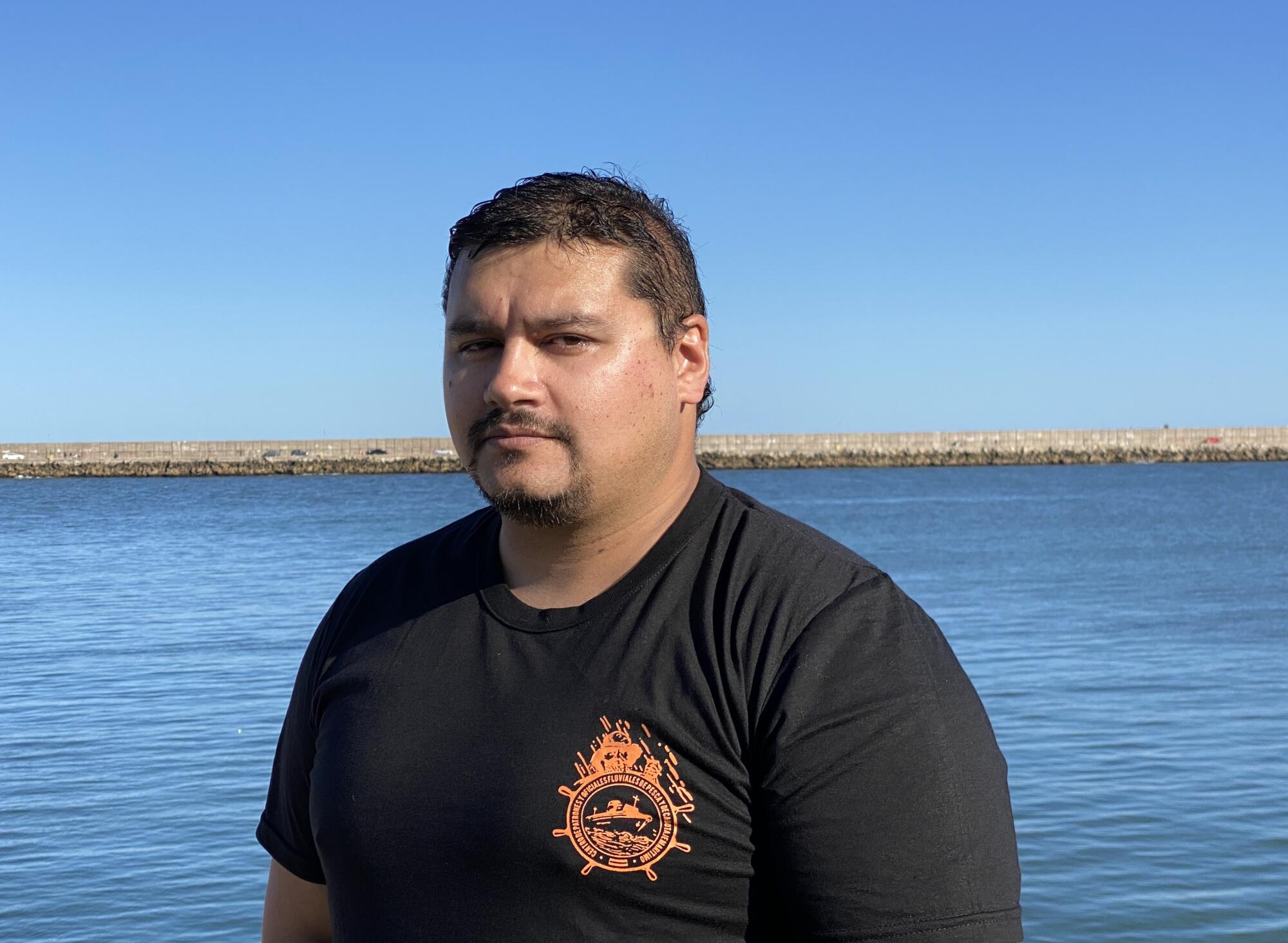 A man with dark hair and mustache, in a dark T-shirt with a wheel logo, with the sea as a backdrop 