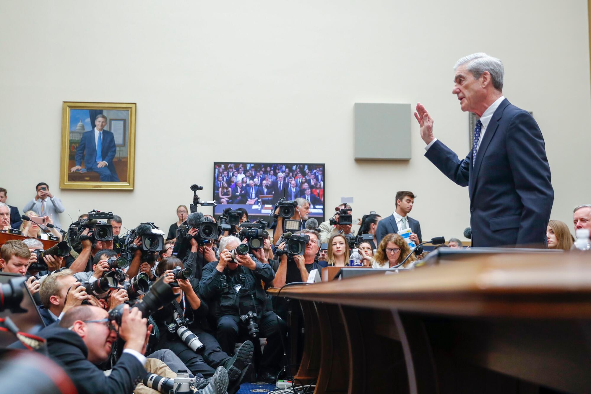 At right Robert Mueller stands behind a desk raising his right hand while facing a crowd of photographers on the ground