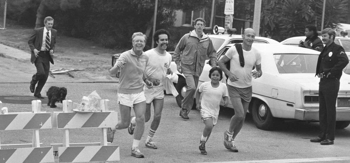 A police officer looks on as five men and a boy jog by on a city street; two men are in street clothes.
