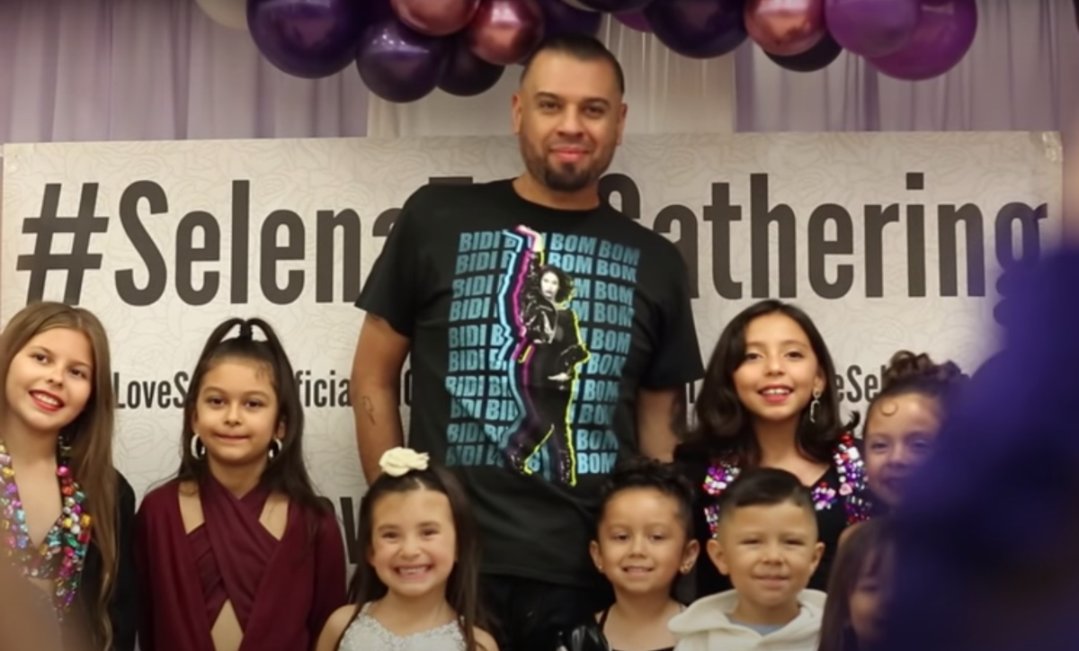 A man in a Selena T-shirt stands with a group of girls in front of a Selena Fan Gathering banner