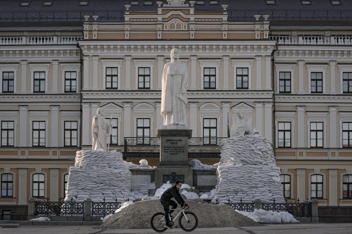 A man rides a bicycle in front of sandbagged statue.