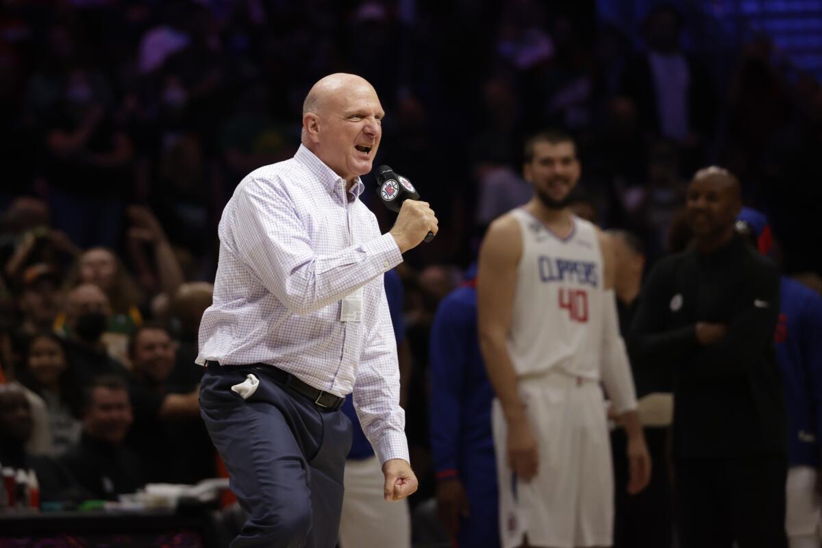 Steve Ballmer speaking into a microphone at a basketball game as a Clippers player and a crowd of fans look on.