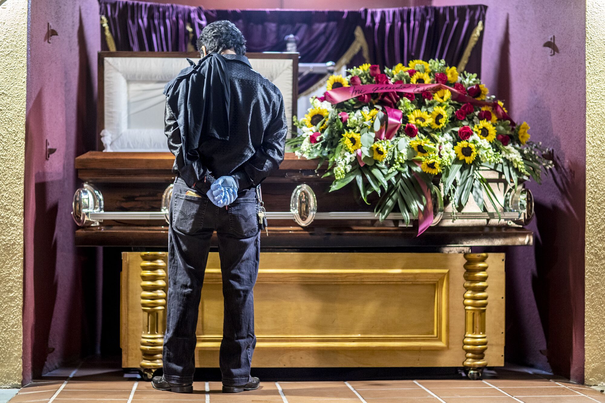 A man stands with hands clasped behind his back in front of a casket covered in sunflowers.