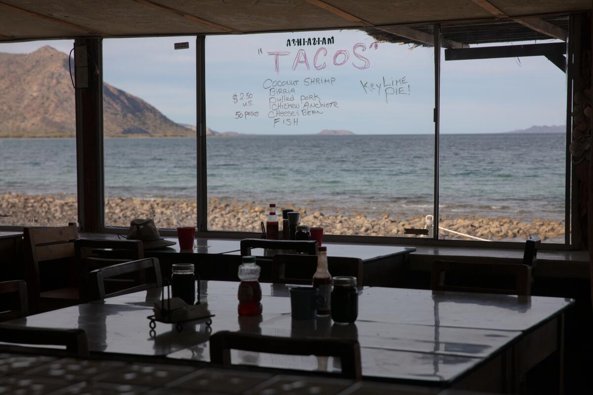 The view out of a window at a pebbly beach; on the window is written "Tacos" and the menu for the restaurant. 