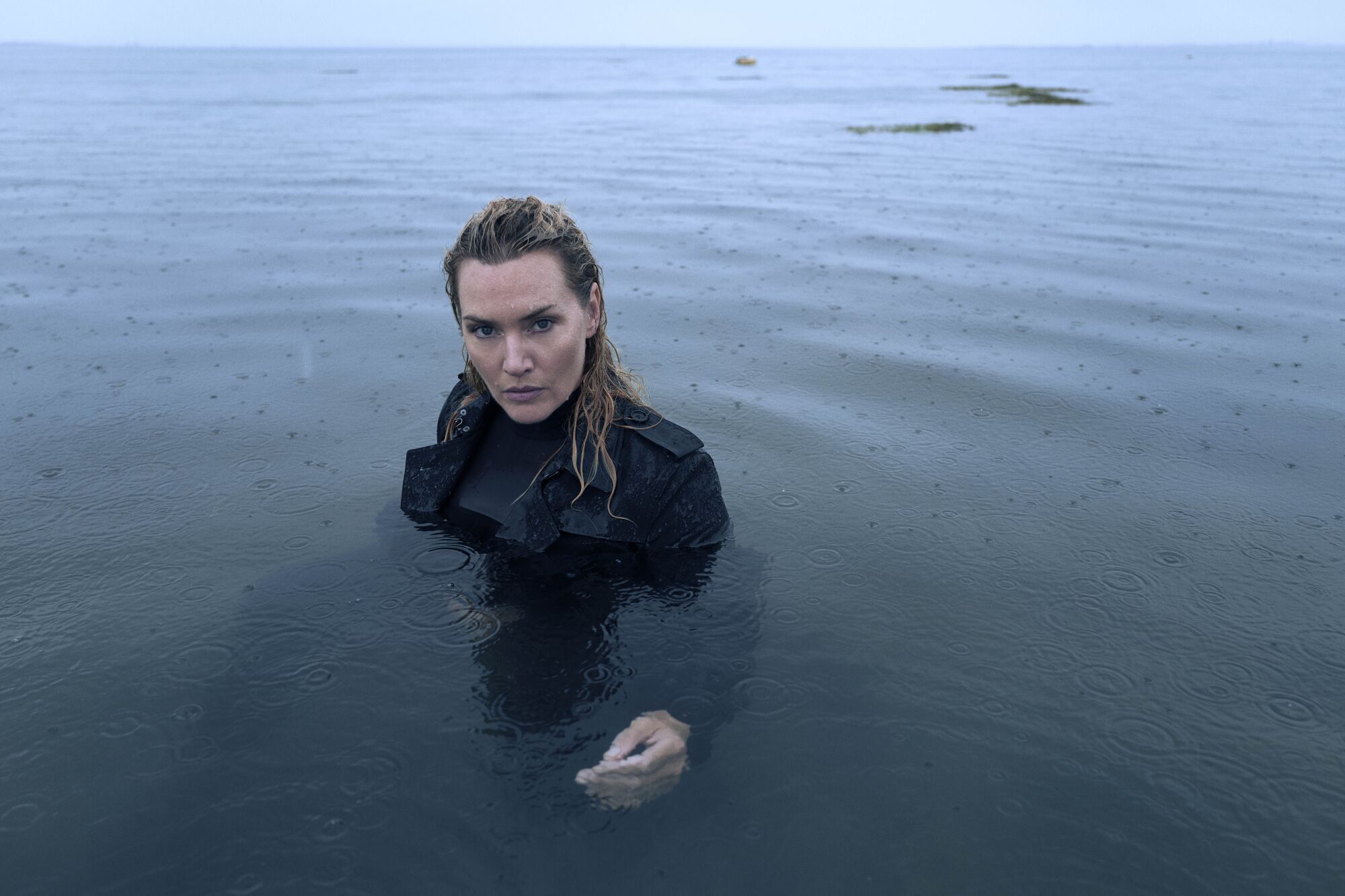 A fully dressed Kate Winslet immerses herself in a body of water in the rain.