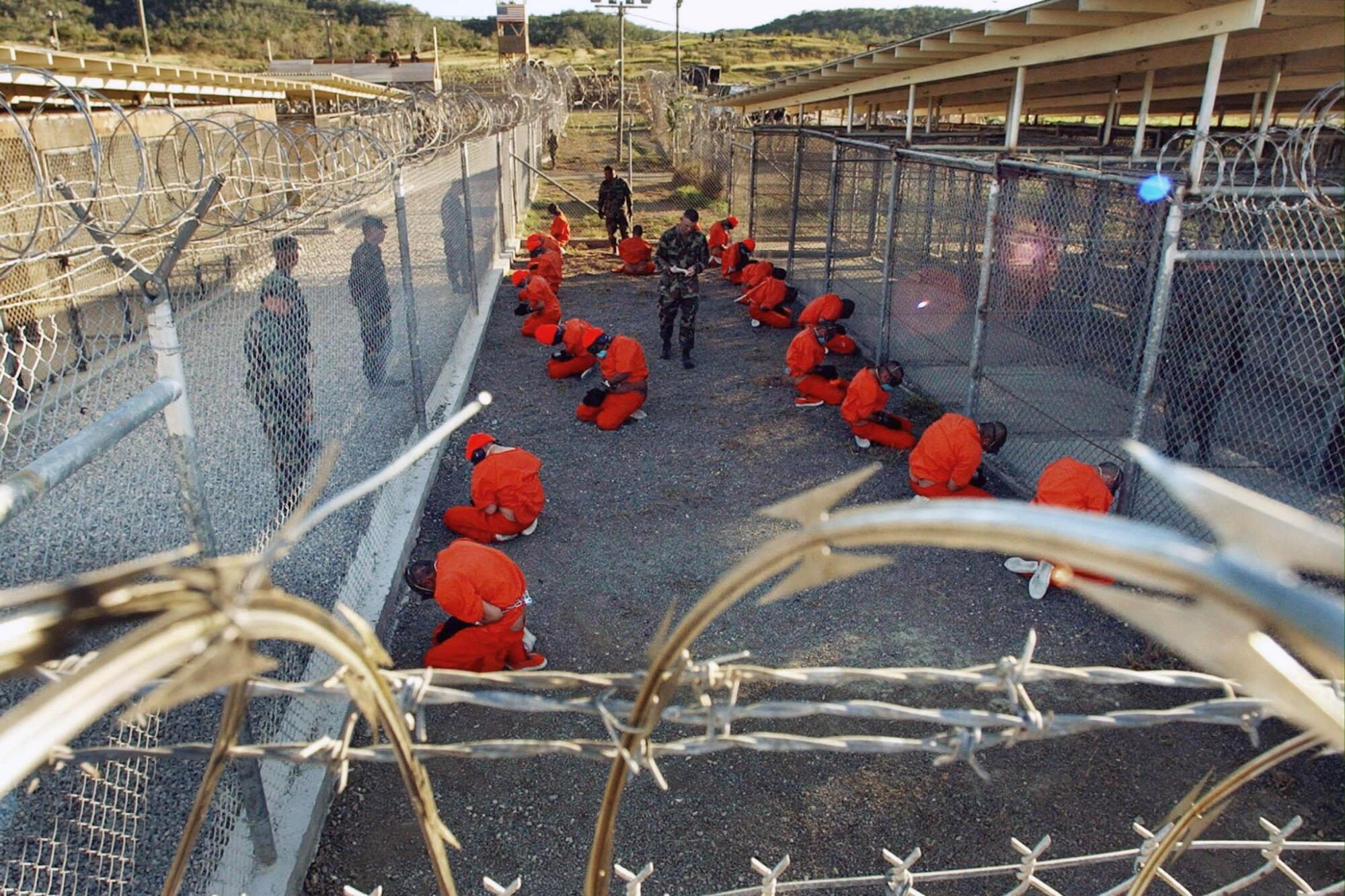 Two rows of men in orange jumpsuits sit in a fenced-off area, watched by men in military fatigues