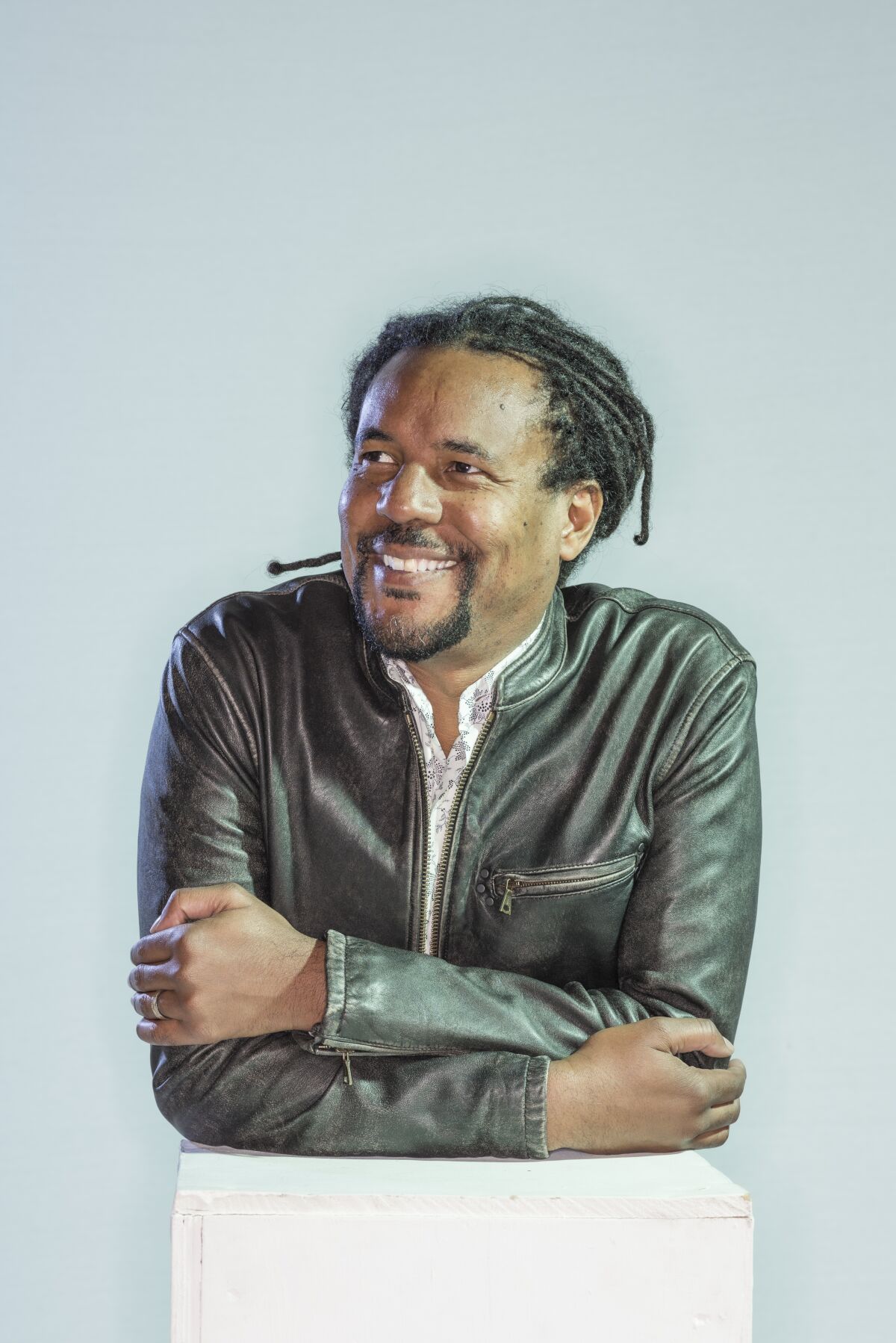 Author Colson Whitehead, wearing a dark jacket and dreadlocks, leans on a white pedestal.