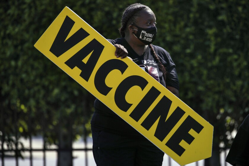 A woman stands outside holding a large sign the reads "Vaccine."