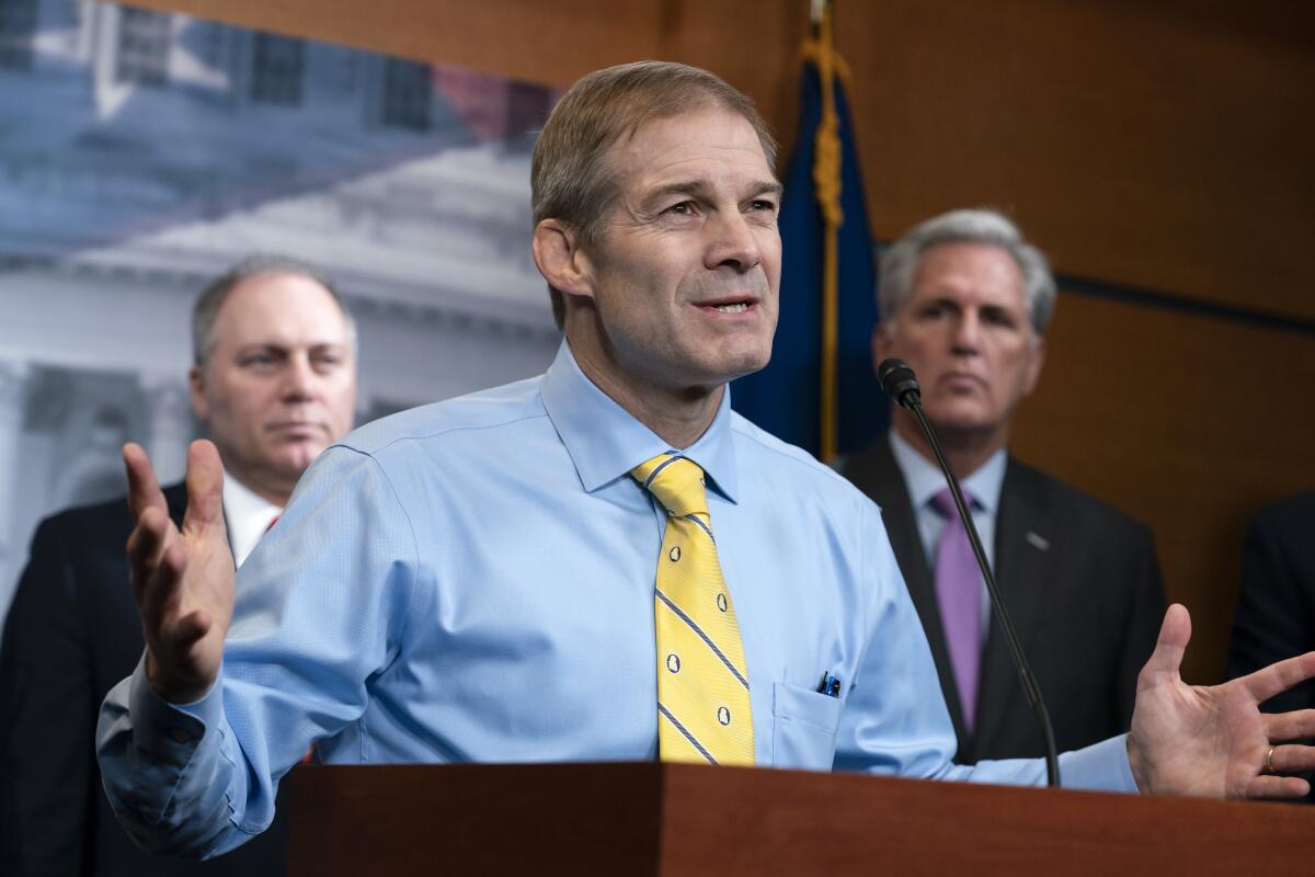 A man in a blue dress shirt, yellow tie and no jacket gestures as he speaks at a lectern