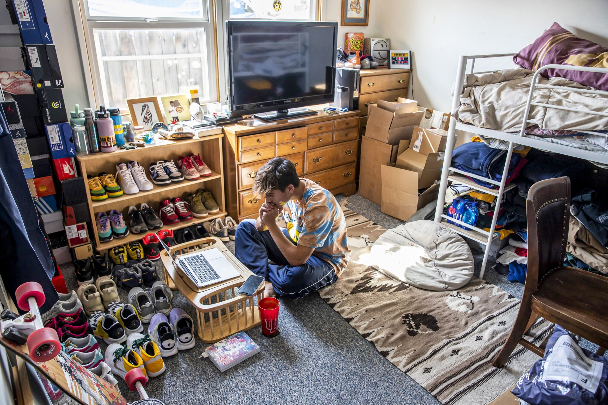 A young man watches a lecture on his computer from the floor of his room filled with shoes and furniture