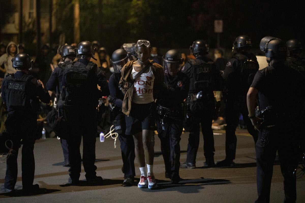 Police officers lead a man with his hands restrained away from a protest scene.