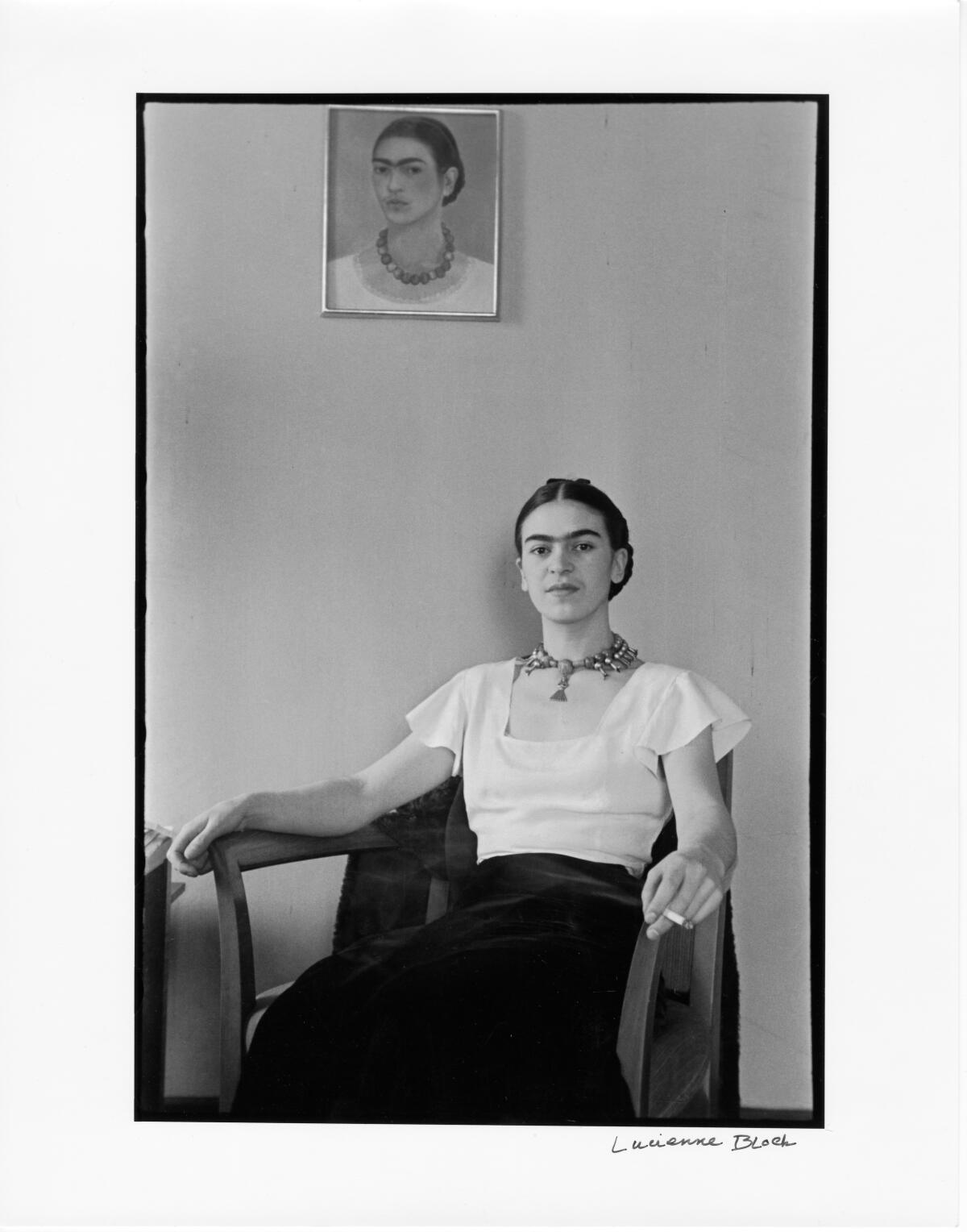 A youthful Frida Kahlo sits smoking in a bare room underneath one of her self-portraits in a vintage black and white photo.