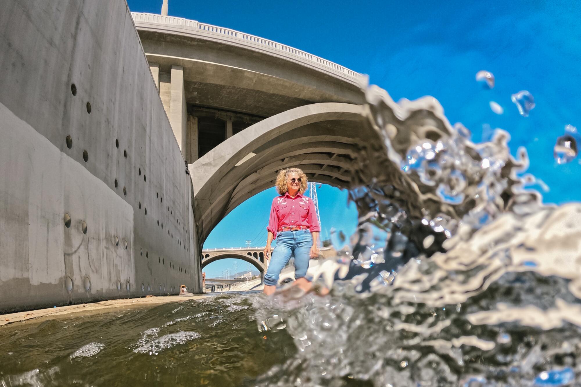 A woman stands inside the Los Angeles river with an overpass behind her.