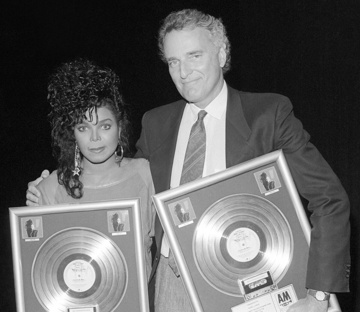 A man stands with his arm around a woman's shoulder. Both hold plaques for gold record album sales