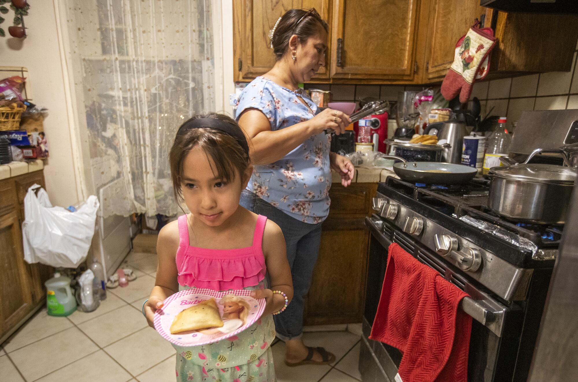 A young girl carries a plate with a quesadilla as her mother cooks in the background