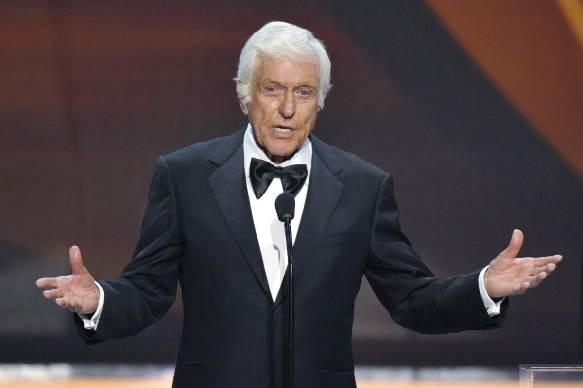 Dick Van Dyke gesturing and speaking into a microphone on a stage while wearing a black suit and bowtie