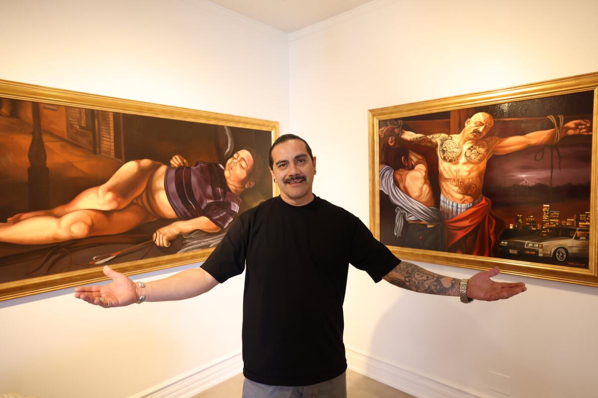 Artist Fabian Debora holds out his arms in front of two paintings, one of someone laying down, the other of a man on a cross
