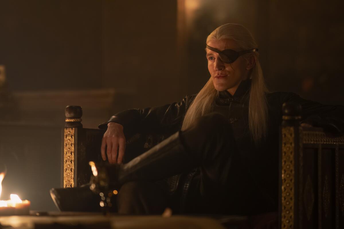 A man with long white hair and an eyepatch sits at a table near a hearth with a fire burning in it