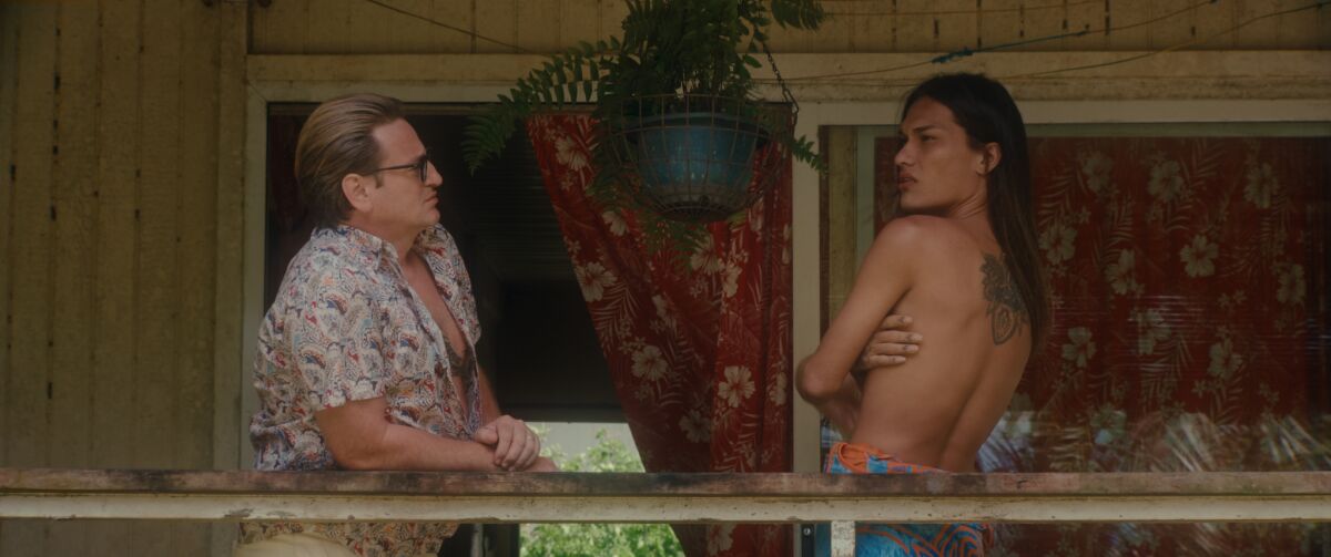 A man in an open aloha shirt faces a topless woman covering her breasts with her arms on a building's porch. 