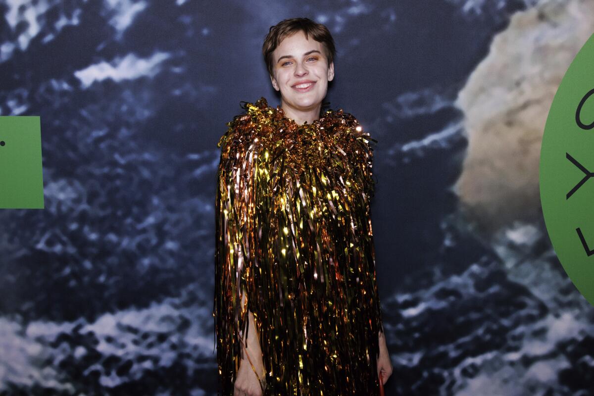 Tallulah Willis wears a shimmering gold dress and smiles as she poses for photos in front of a blue and white background.