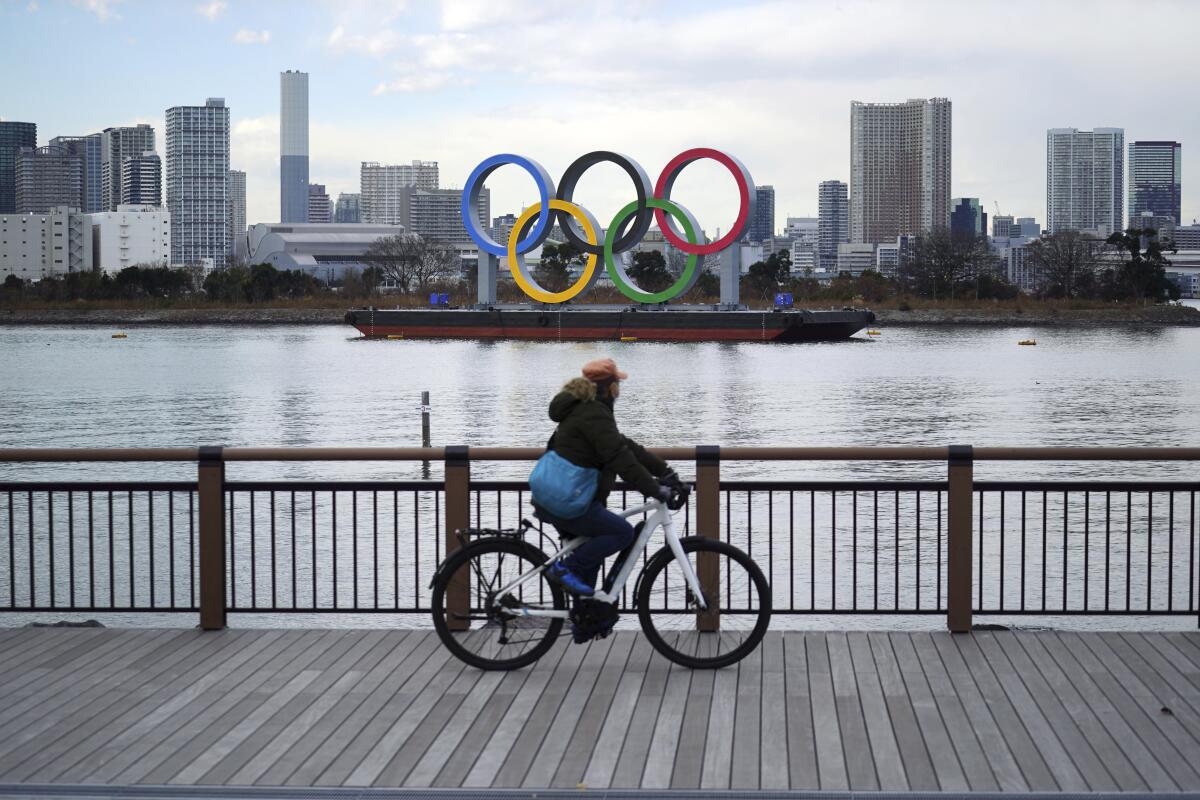 Olympic rings float in the water on a boat while a cyclist bikes by on a boardwalk