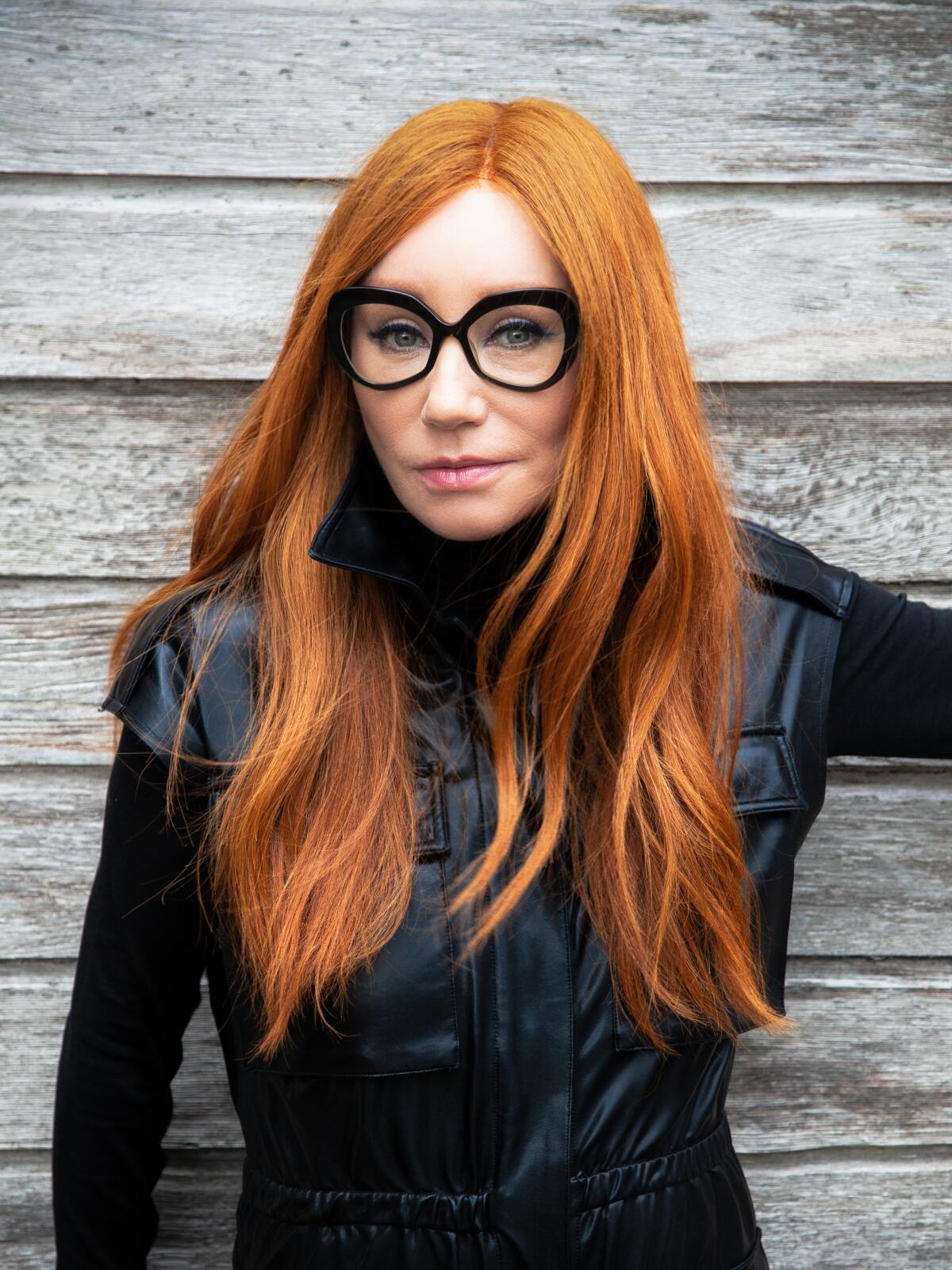 A woman with long red hair, black-framed glasses and a black leather jacket.