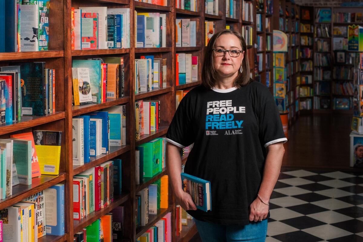 A dark haired woman in glasses and a dark colored shirt holds a book next to shelves full of books. 