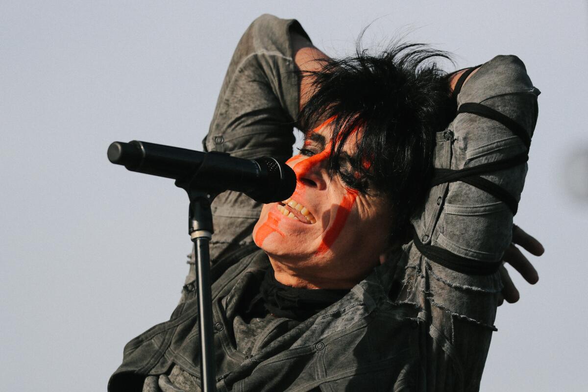 A man at a microphone with dyed black hair and streaky orange facepaint stands with his arms behind his head.