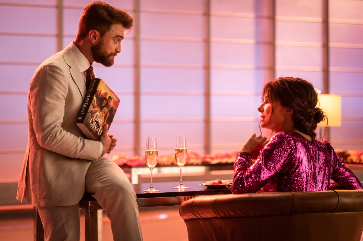 A man in a suit leans against a table holding up a book to a woman in a pink sequined dress in a chair 