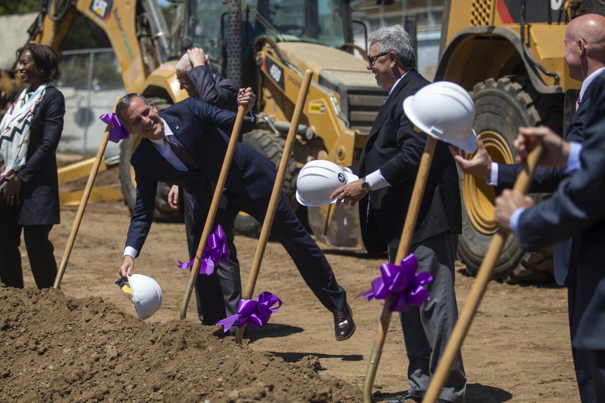 Eric Garcetti grasps the handle of a shovel, one among several stuck in a mound of dirt and decorated with purple ribbon.