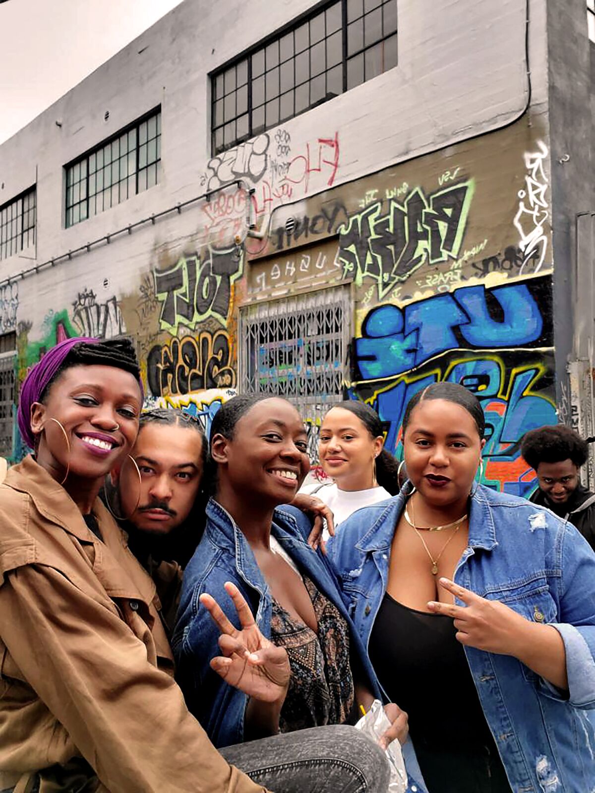 Six young adults smiling outside a building covered with graffiti. 