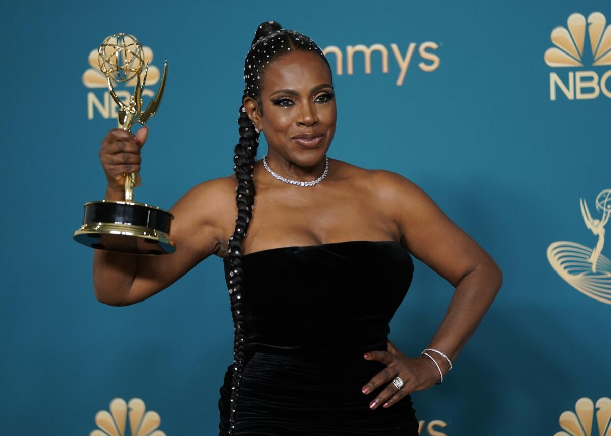 A woman in a black strapless dress and long braided hair poses for pictures while holding her Emmy award.