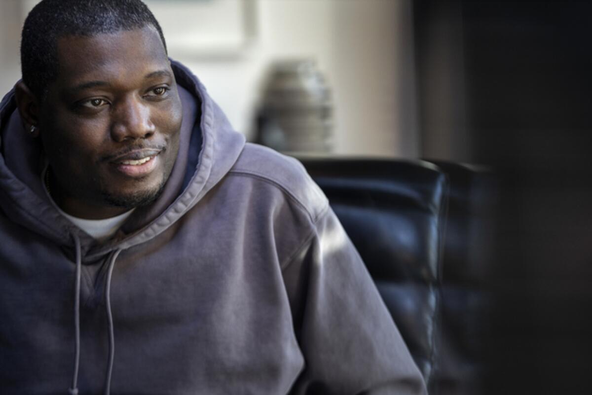Michael Che sits in a chair while wearing a hoodie with a white T-shirt peeking from the collar.