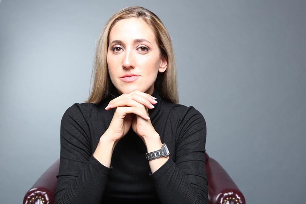 A blond woman, seated, resting her chin on her clasped hands.