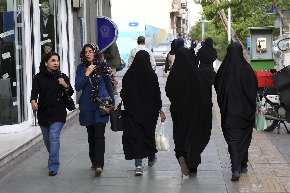 Iranian women walk on a sidewalk. Several are in black outfits that cover them from head to foot. Two wear street clothes.