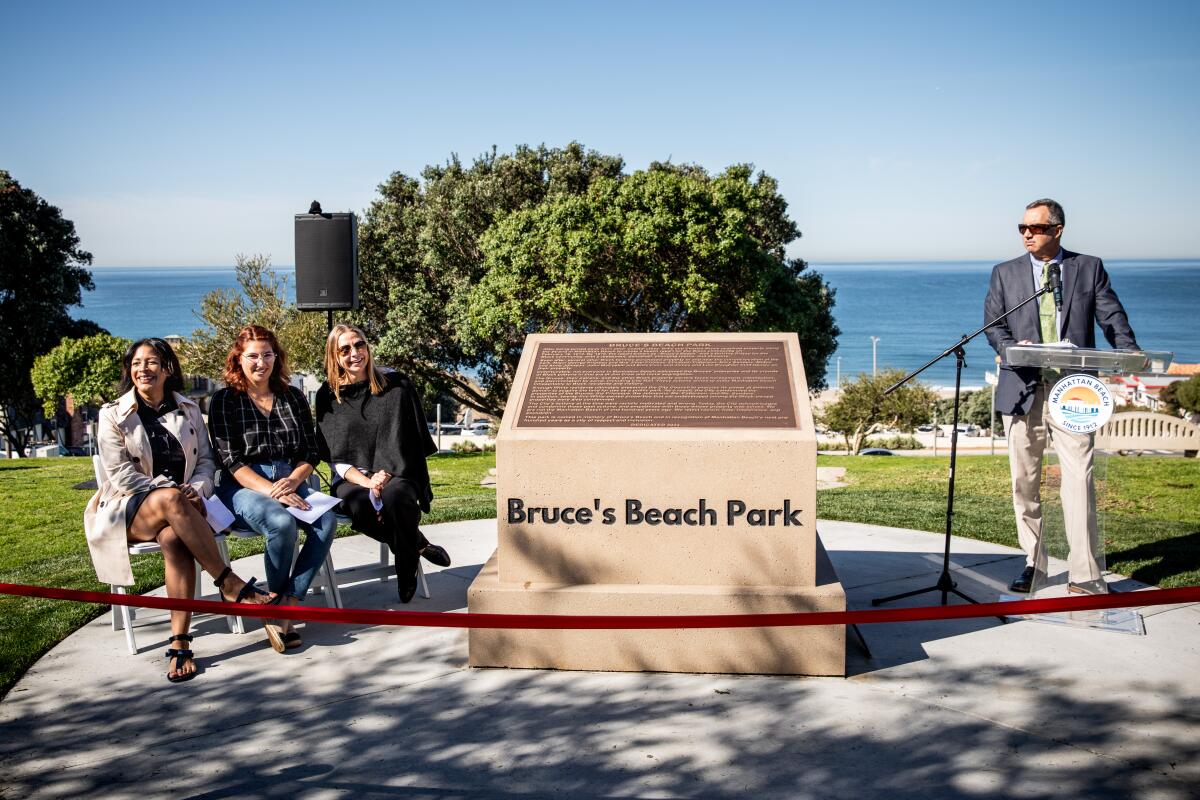 Three people sit on a bench next to a plaque as a man stands at a microphone