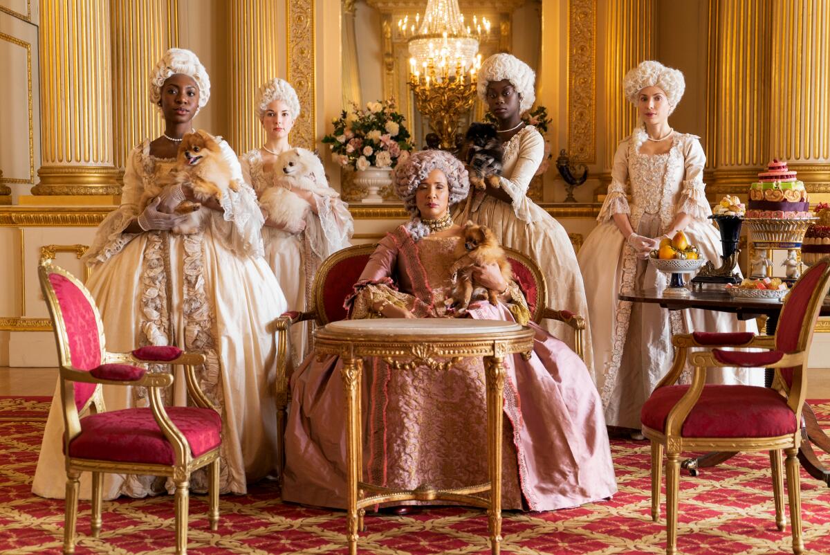 In a room with gilded furnishings, five women wear elaborate Regency gowns and wigs and carry Pomeranians.