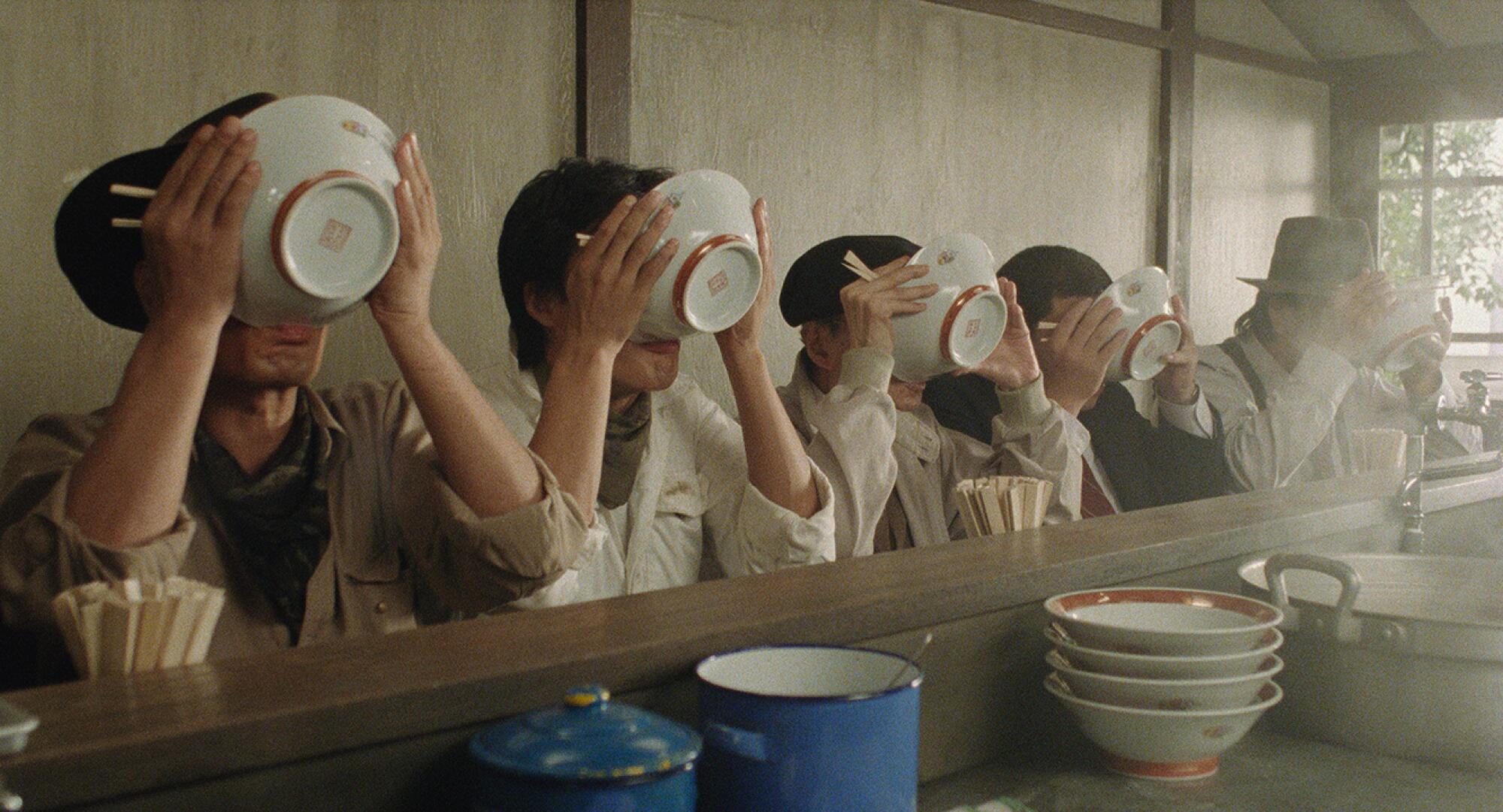 A line of customers at the counter of a ramen restaurant hold bowls of soup up to their mouths.