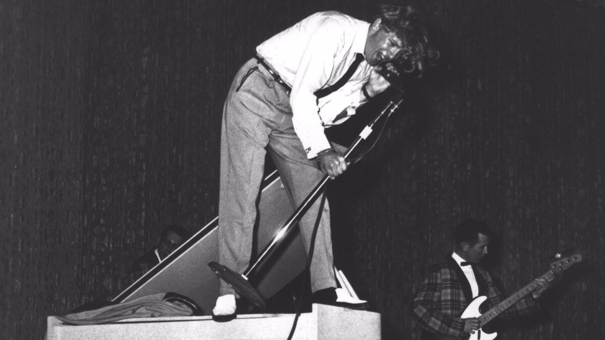 Jerry Lee Lewis stands atop a piano, holding a microphone stand.
