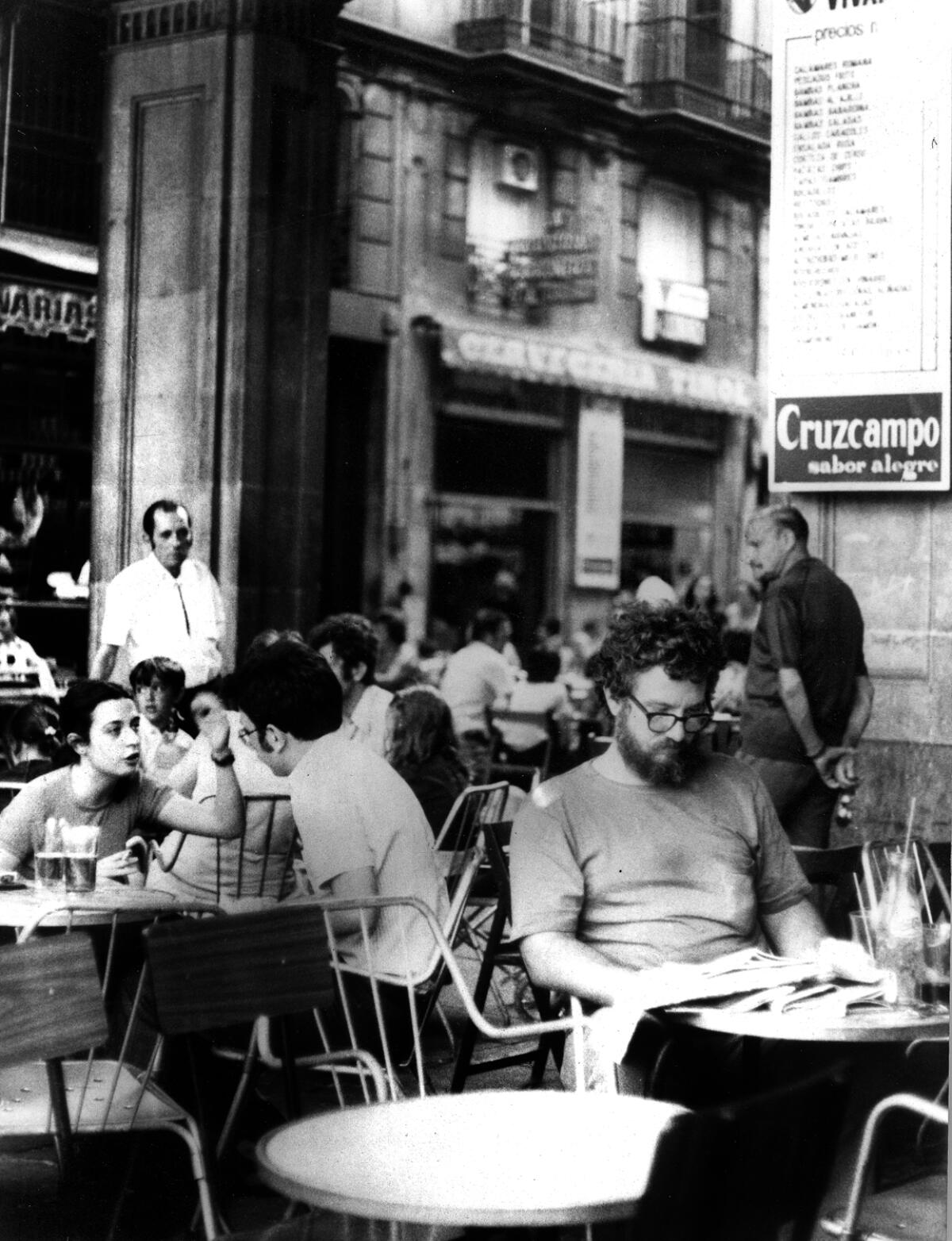 A bearded young man sits at an outdoor cafe table