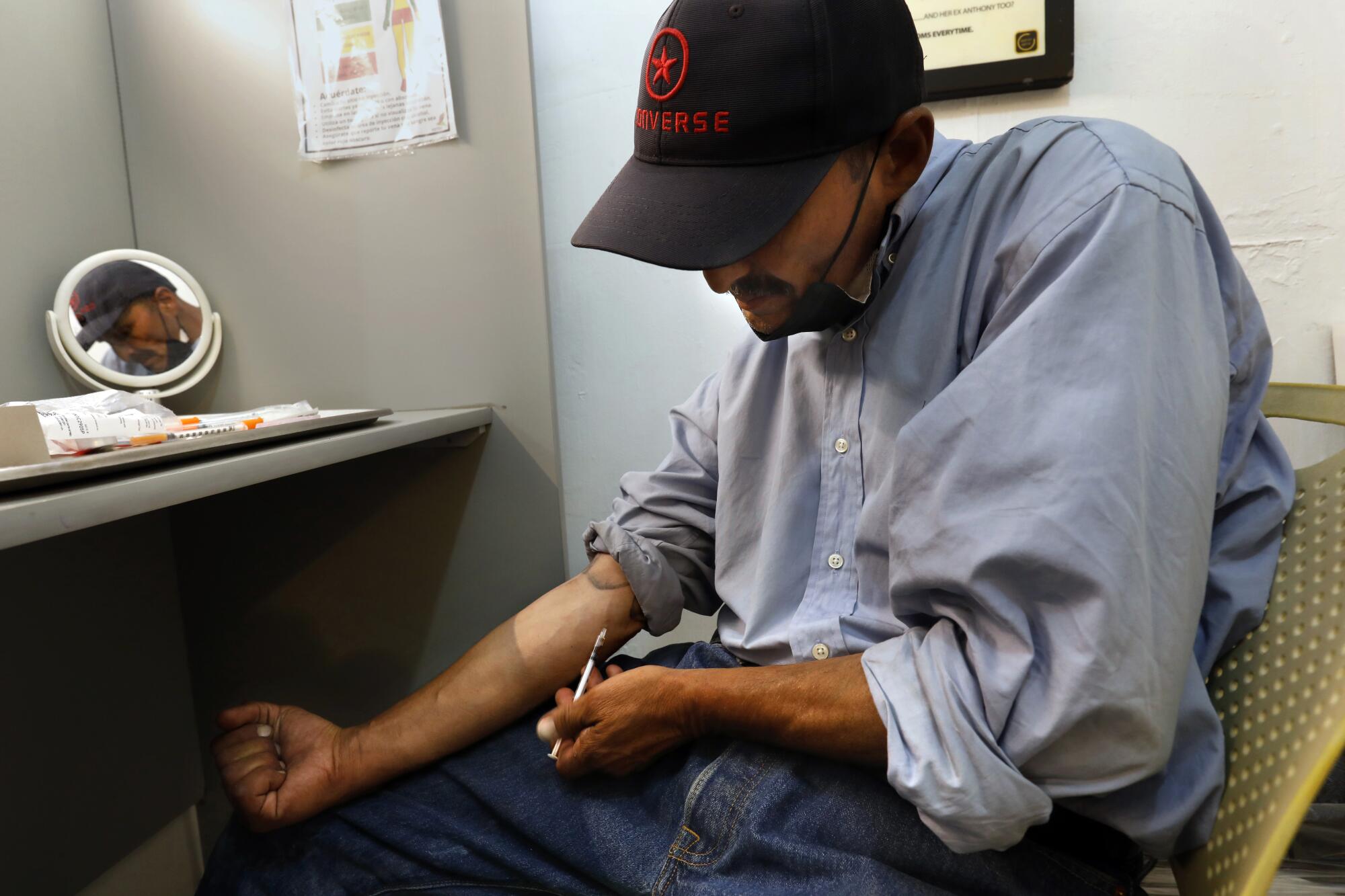 A man, his face obscured by a cap, holds a needle to the crook of his arm while seated 