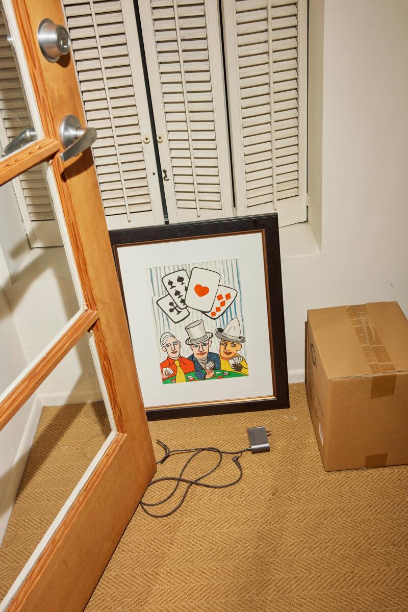 A framed Alexander Calder lithograph of three card players sits on the wood floor of a room.