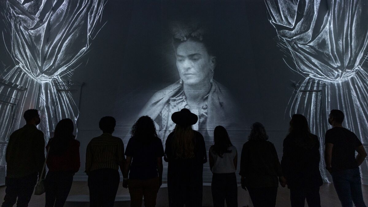 People are seen in silhouette standing in front of a large black and white portrait of Frida Kahlo projected on a wall.