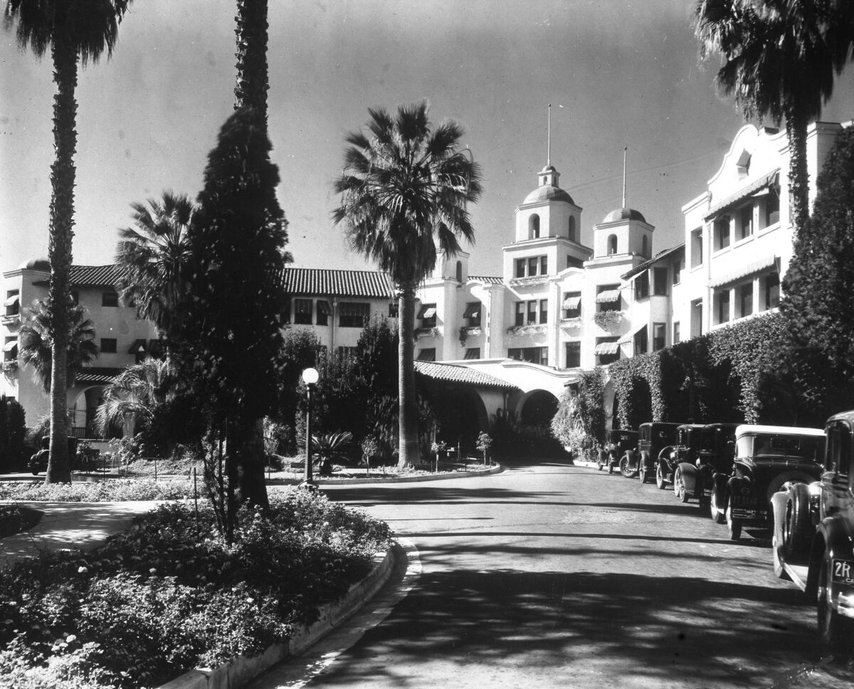 A multistory white building built in a curve, with mature palm trees and Model T-era cars parked along a driveway.