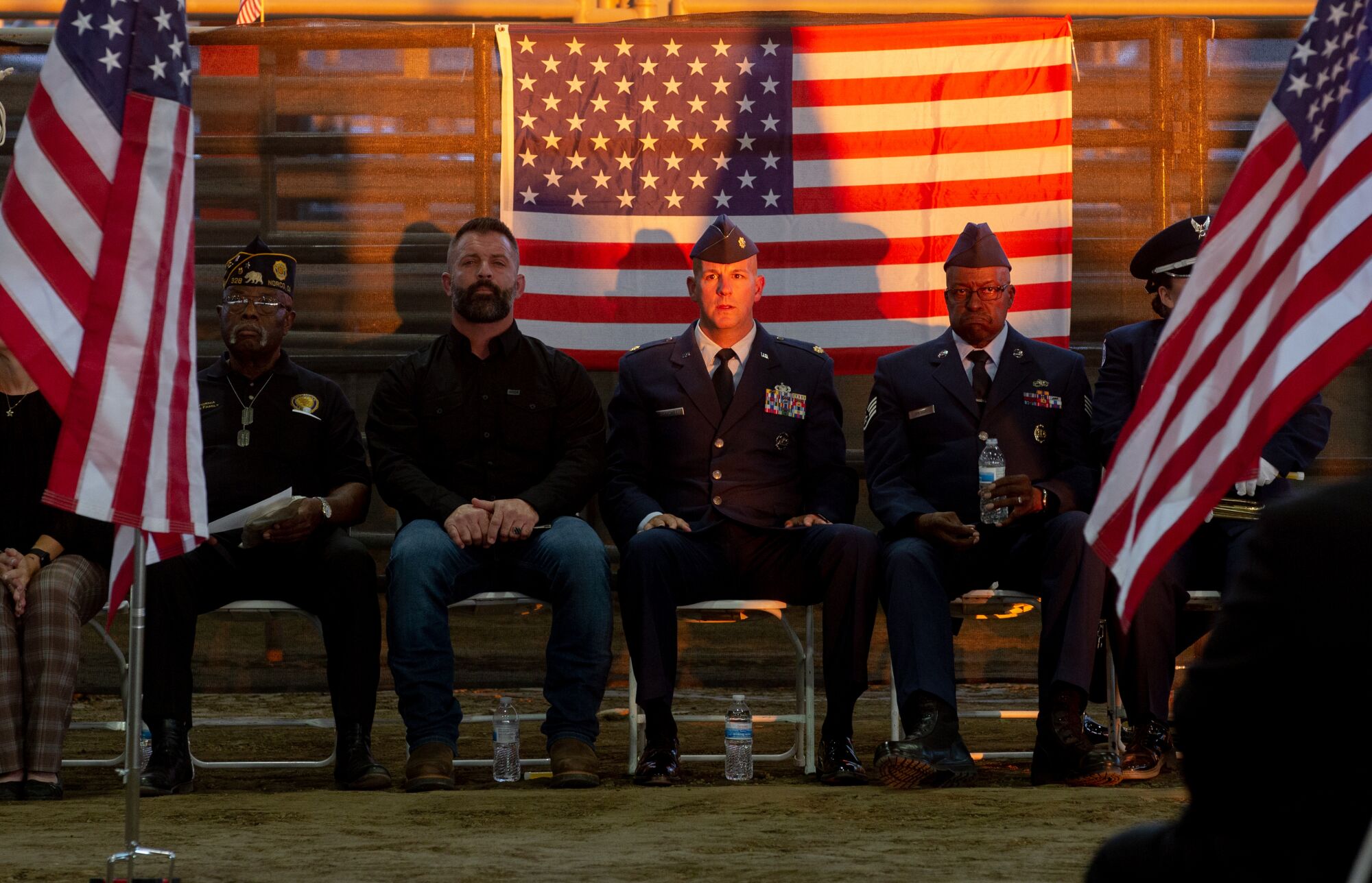 Men sit in chairs in front of a U.S. flag. Some are in military uniform.