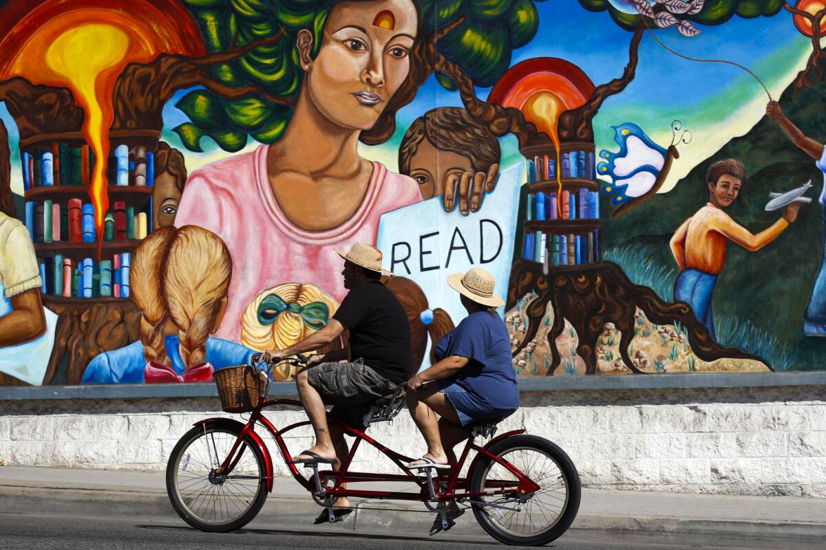 An image of two people riding a bike past a colorful mural that shows a woman holding a sign that says "Read"