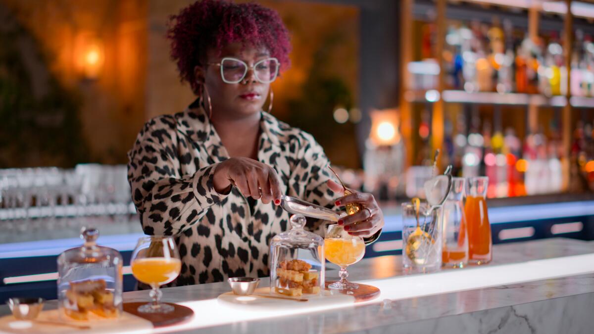 A woman bartender in leopard print shirt, statement glasses and hoop earrings mixes an elaborate cocktail