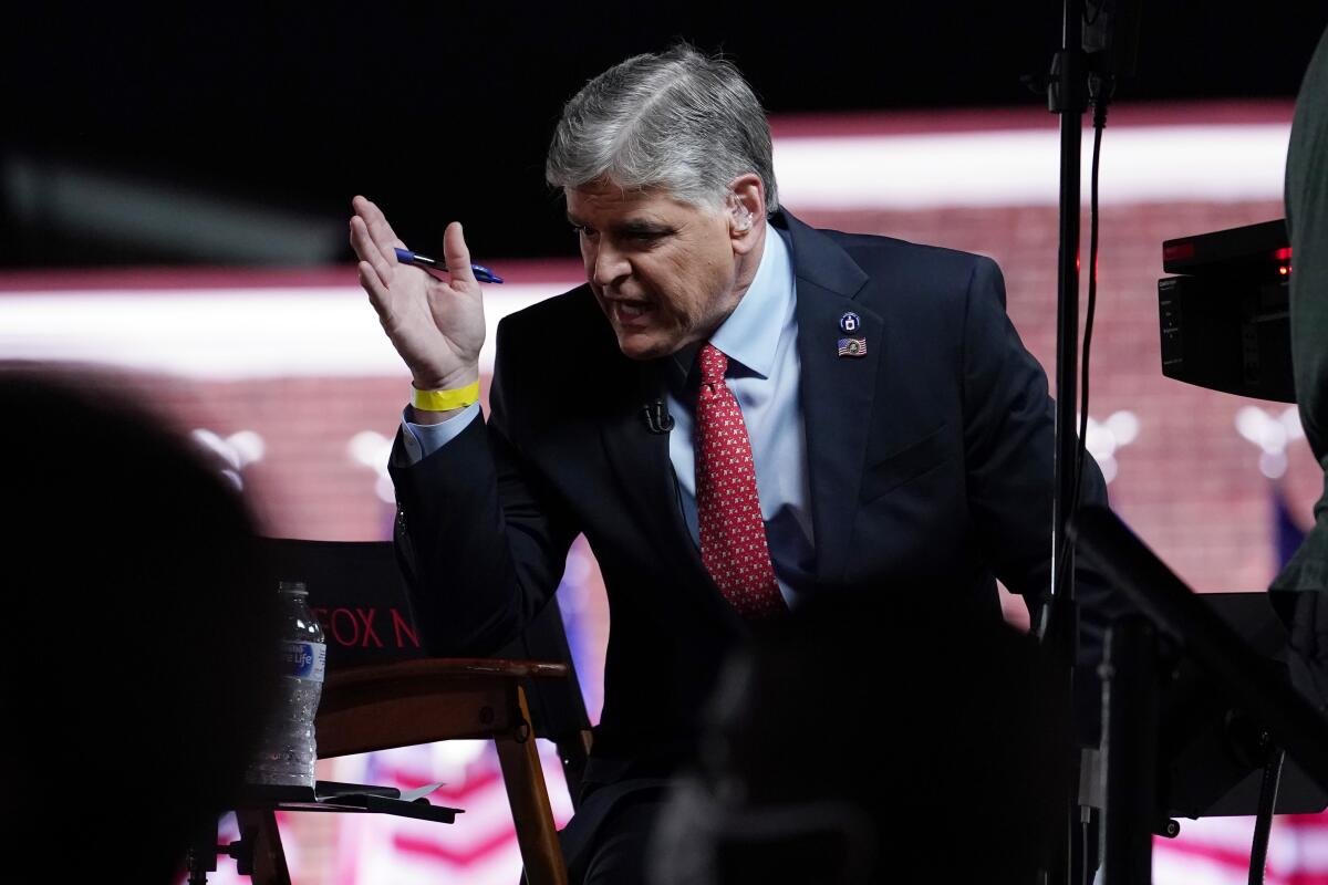 Sean Hannity, in suit and tie, sits on a stage and gestures emphatically.