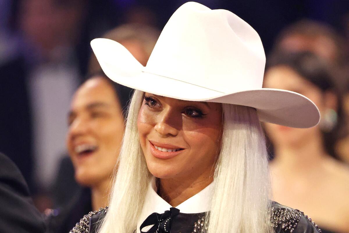 Beyoncé with long platinum hair, wearing a wide-brimmed hat, smiling and tilting her head